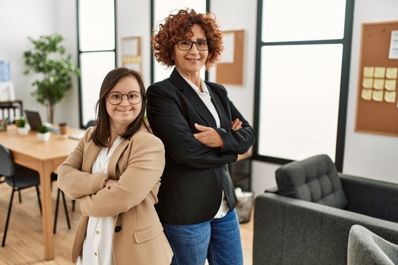 Eine Frau in mittlerem Alter und eine junge Frau mit Downsyndrom stehen nebeneinander in einem Büro für Leichte Sprache. Im Hintergrund ist ein Arbeitsplatz mit Laptop zu sehen.