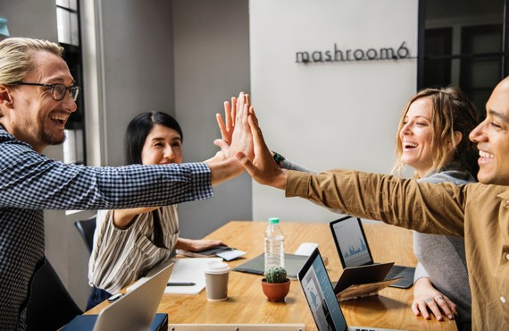 Zwei Frauen und zwei Männer in einer Agentur oder in einem Unternehmen sitzen zusammen an einem Besprechungstisch. Sie freuen sich und geben sich High Five.