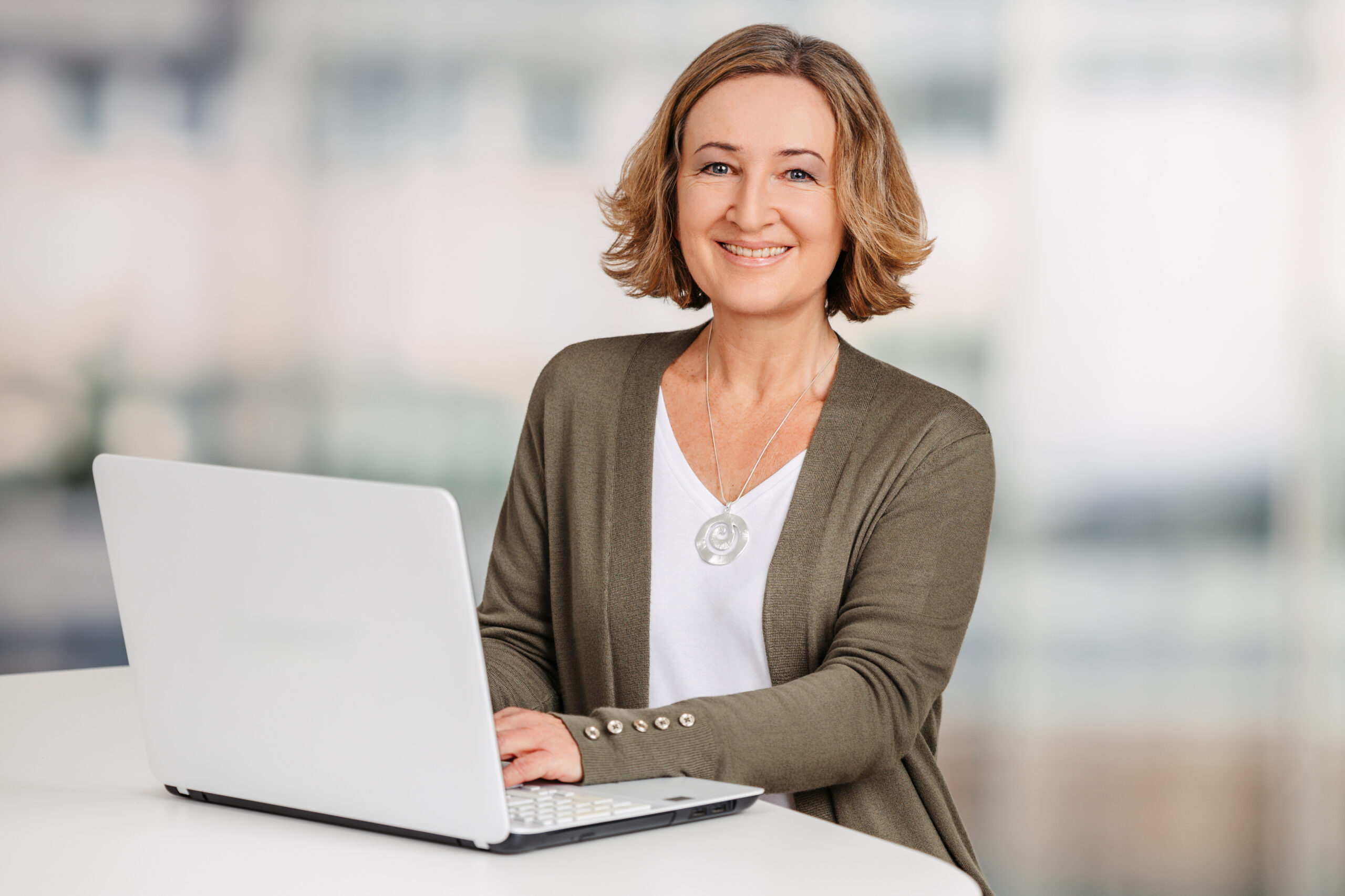 Petra Eppig steht an einem hohen weißen Tisch und arbeitet am Laptop. Petra Eppig hat halblange, dunkelblonde Haare und trägt ein weißes Shirt, eine dunkelgrüne Strickjacke und eine Kette mit einem silbernen Anhänger. Sie lächelt in die Kamera.