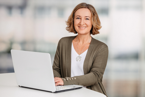 Petra Eppig steht an einem hohen weißen Tisch und arbeitet am Laptop. Petra Eppig hat halblange, dunkelblonde Haare und trägt ein weißes Shirt, eine dunkelgrüne Strickjacke und eine Kette mit einem silbernen Anhänger.