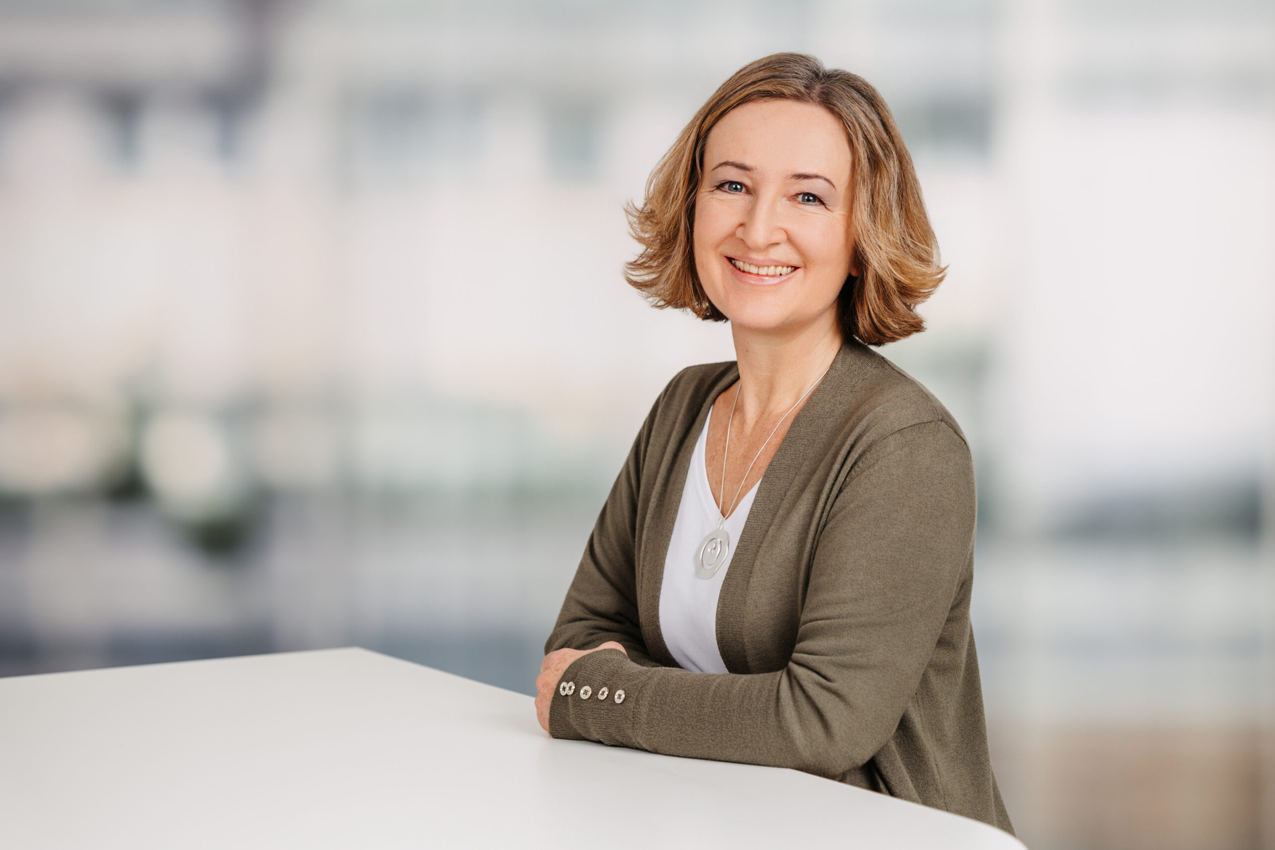 Ein Foto von Petra Eppig. Petra Eppig ist Lektorin und Übersetzerin für Leichte Sprache und Einfache Sprache. Sie steht an einem hohen weißen Tisch. Petra Eppig hat halblange, dunkelblonde Haare und trägt ein weißes Shirt, eine dunkelgrüne Strickjacke und eine Kette mit einem silbernen Anhänger. Sie lächelt in die Kamera.