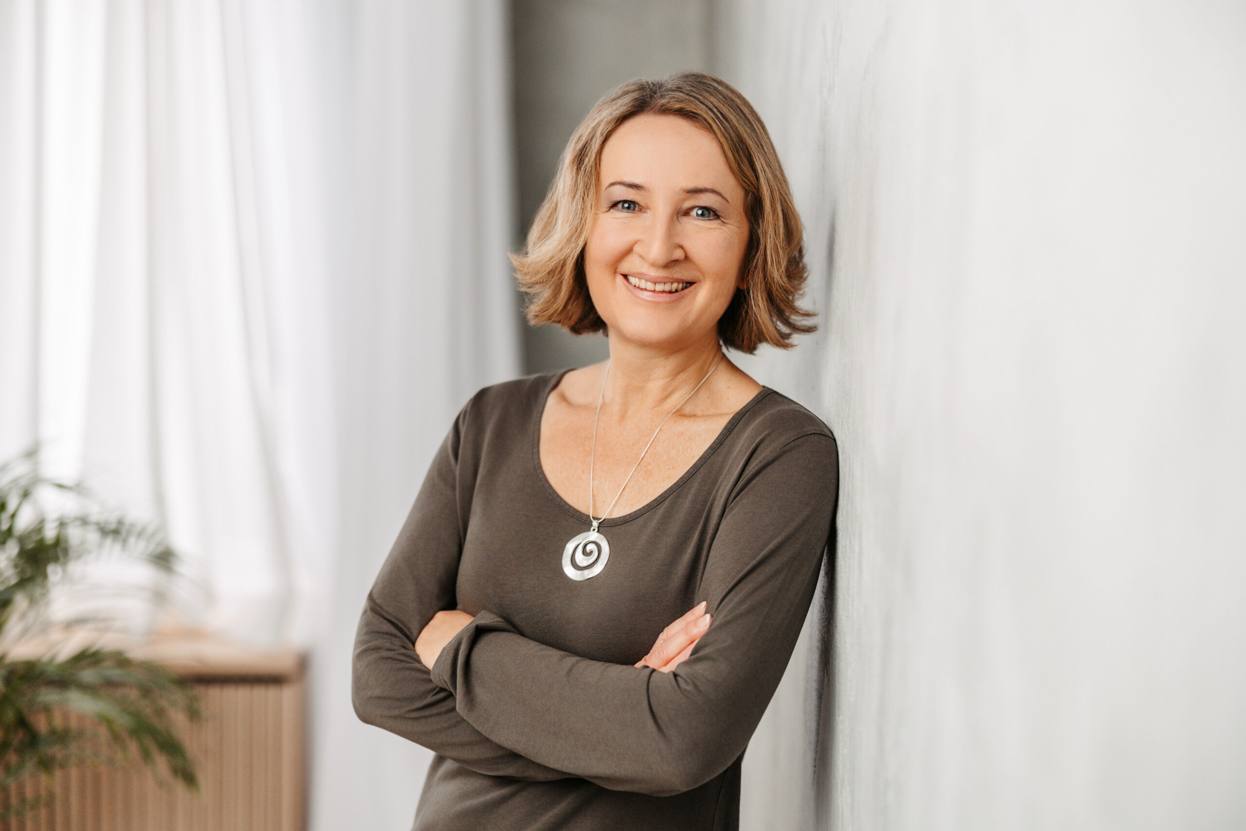 Ein Foto von Petra Eppig. Petra Eppig ist Lektorin und Übersetzerin für Leichte Sprache und Einfache Sprache. Sie lehnt sich mit der Schulter an eine helle Wand. Petra Eppig hat halblange, dunkelblonde Haare und trägt ein braunes Shirt und eine Kette mit einem silbernen Anhänger. Sie lächelt in die Kamera.